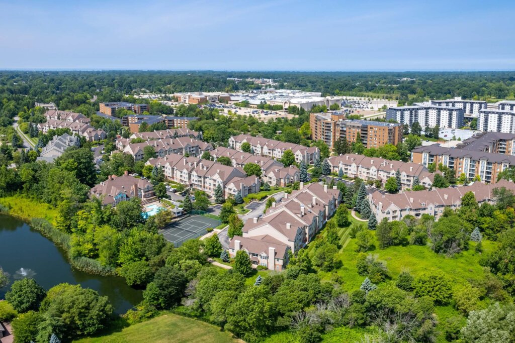 Aerial view of a residential community in Brookfield, Wisconsin, featuring townhouses, apartment buildings, lush greenery, a pond, and tennis courts, with a shopping mall in the background.