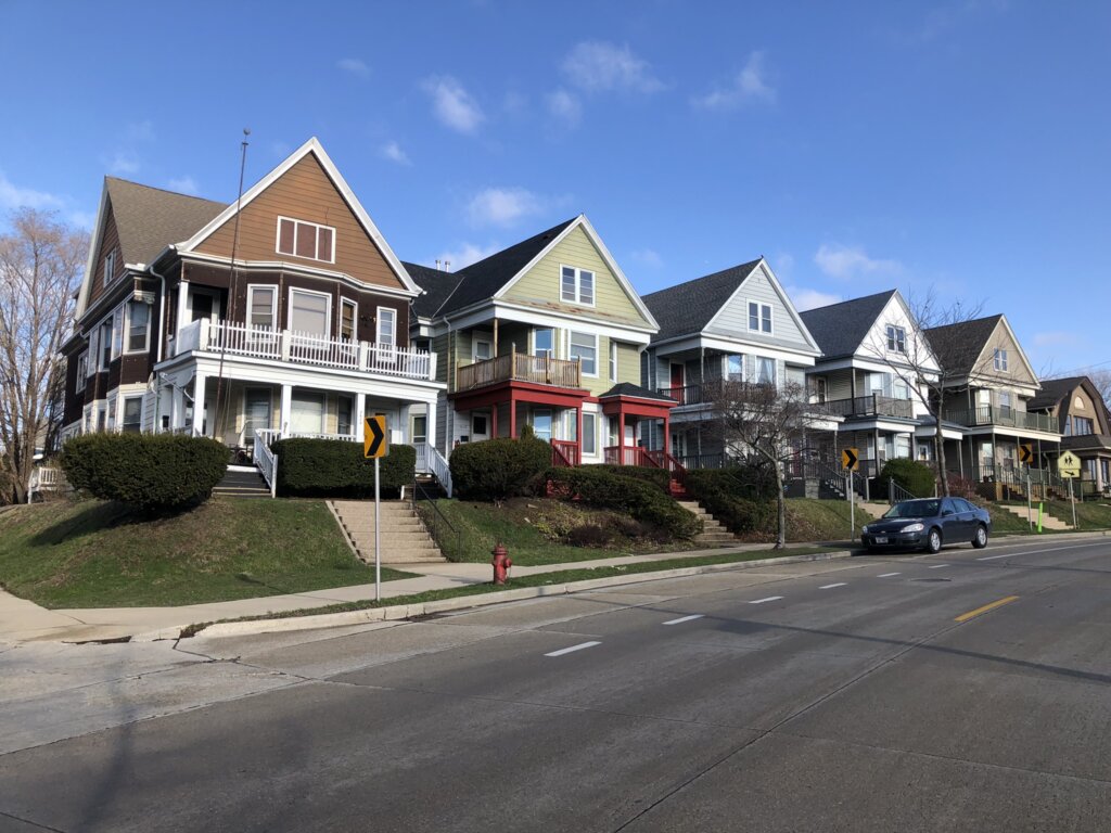 A row of colorful, well-maintained houses with gabled roofs along a sloped street in Milwaukee, featuring porches, staircases, and a parked car under a clear blue sky.