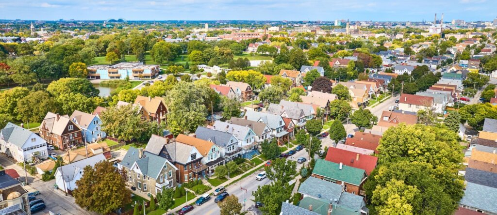 Aerial view of a Milwaukee neighborhood with colorful houses, tree-lined streets, and a large park in the background on a sunny day.
