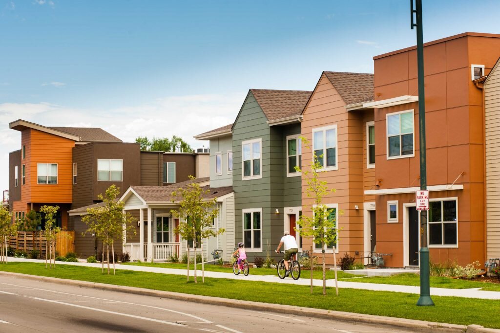 Modern row of colorful townhouses along a green suburban street, with a cyclist and a child riding a bike on the sidewalk.