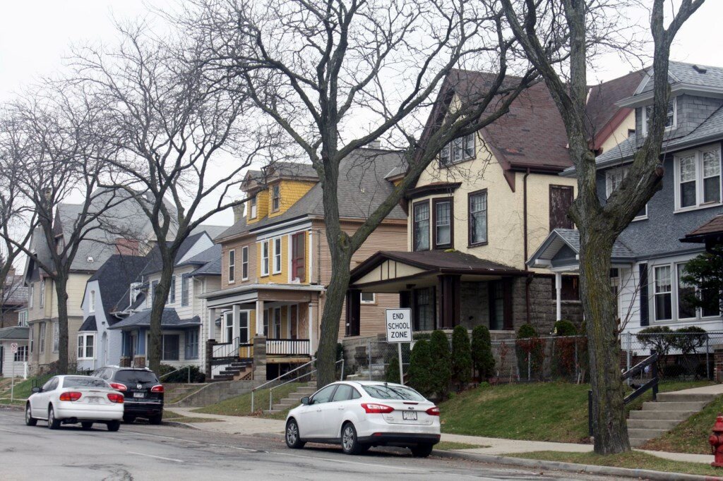 A row of historic houses on a residential street in Milwaukee, with leafless trees lining the sidewalk and parked cars along the curb.