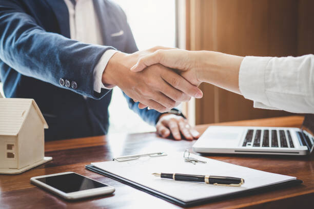 Close-up of a handshake between two people finalizing a real estate deal, with a contract, pen, and house model on the table.