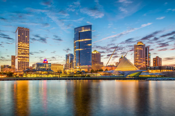 A vibrant view of downtown Milwaukee, Wisconsin, at sunset. The city's skyline features modern skyscrapers reflecting on the water, with the striking architecture of the Milwaukee Art Museum visible in the foreground. The warm orange and blue hues of the sky add to the scenic beauty.