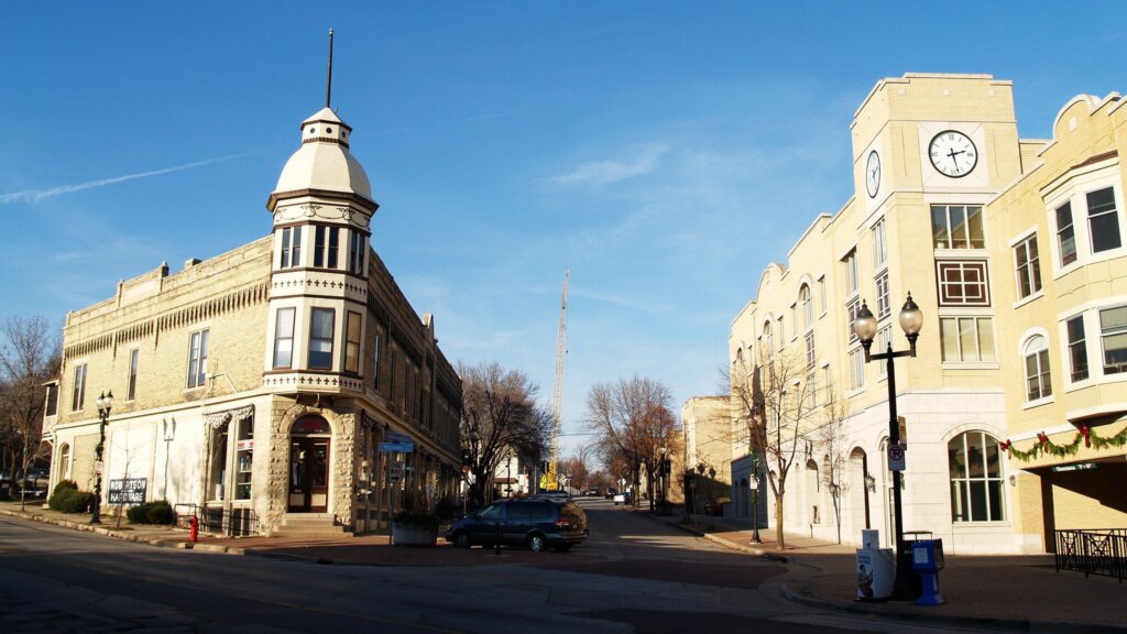 The image shows a street in a small town with historic and modern buildings, including a clock tower on one side and a construction crane in the background.