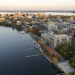 An aerial view of Madison, Wisconsin, showing the city skyline, Lake Mendota, and Helen C. White Hall, with vibrant autumn foliage.