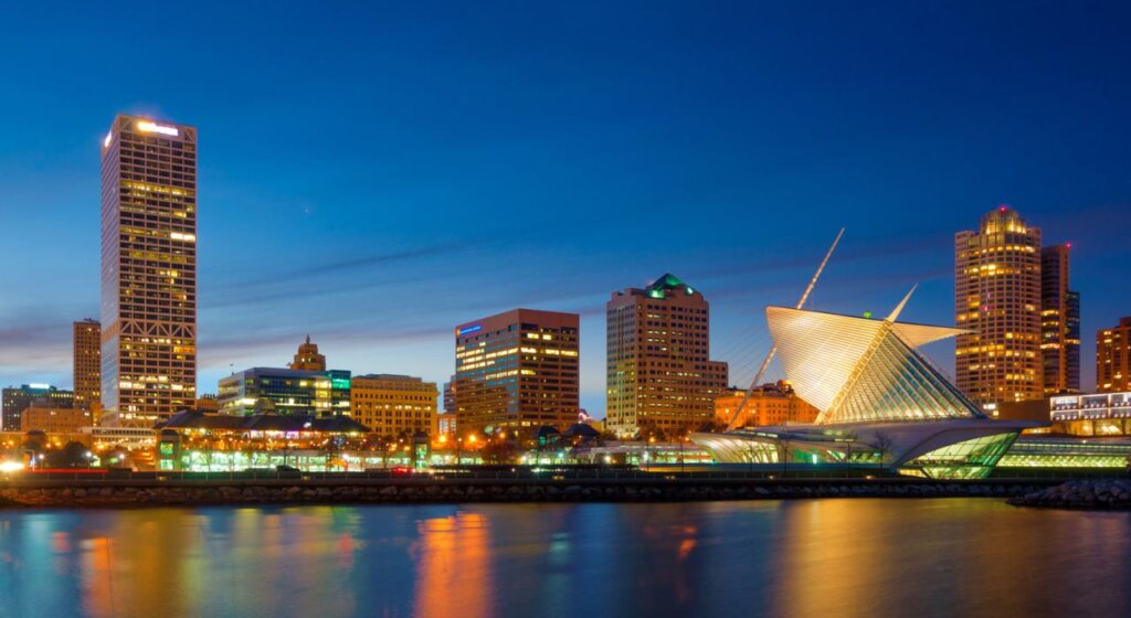 Milwaukee skyline at dusk featuring the illuminated Milwaukee Art Museum and surrounding city buildings reflected on Lake Michigan.