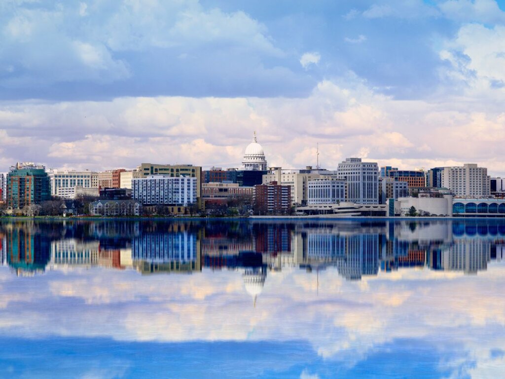 Downtown Madison skyline with the Wisconsin State Capitol reflected in Lake Monona under a partly cloudy sky.
