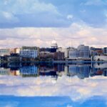 Downtown Madison skyline with the Wisconsin State Capitol reflected in Lake Monona under a partly cloudy sky.