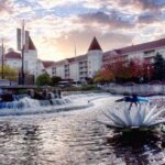 Scenic view of a river with a waterfall and sculpture in front of a large castle-like building at sunset in Waukesha.