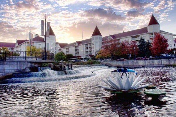 Scenic view of a river with a waterfall and sculpture in front of a large castle-like building at sunset in Waukesha.