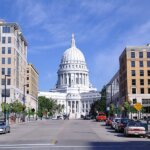 View of the Wisconsin State Capitol building in Madison, framed by modern office buildings and parked cars on a sunny day.