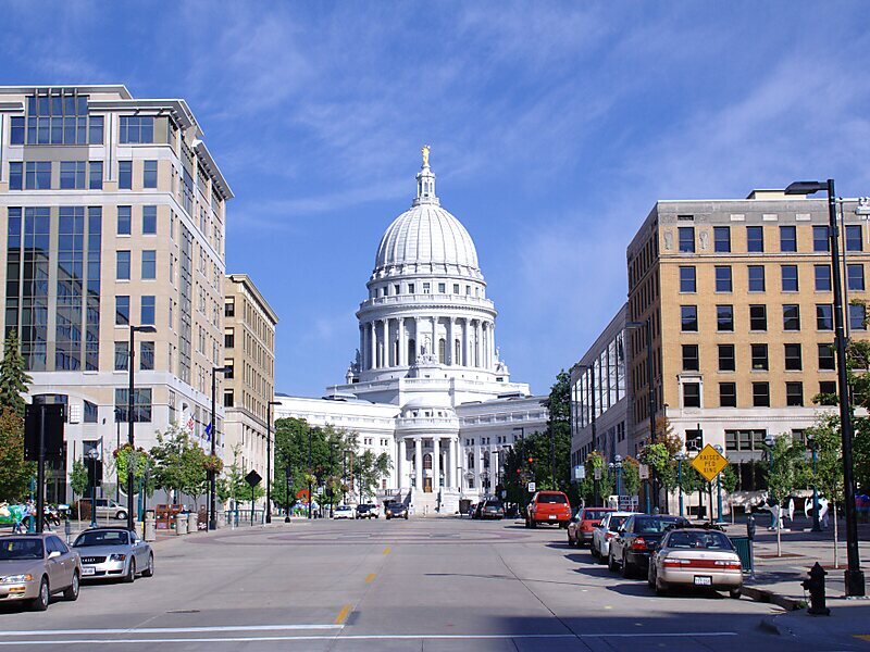 View of the Wisconsin State Capitol building in Madison, framed by modern office buildings and parked cars on a sunny day.
