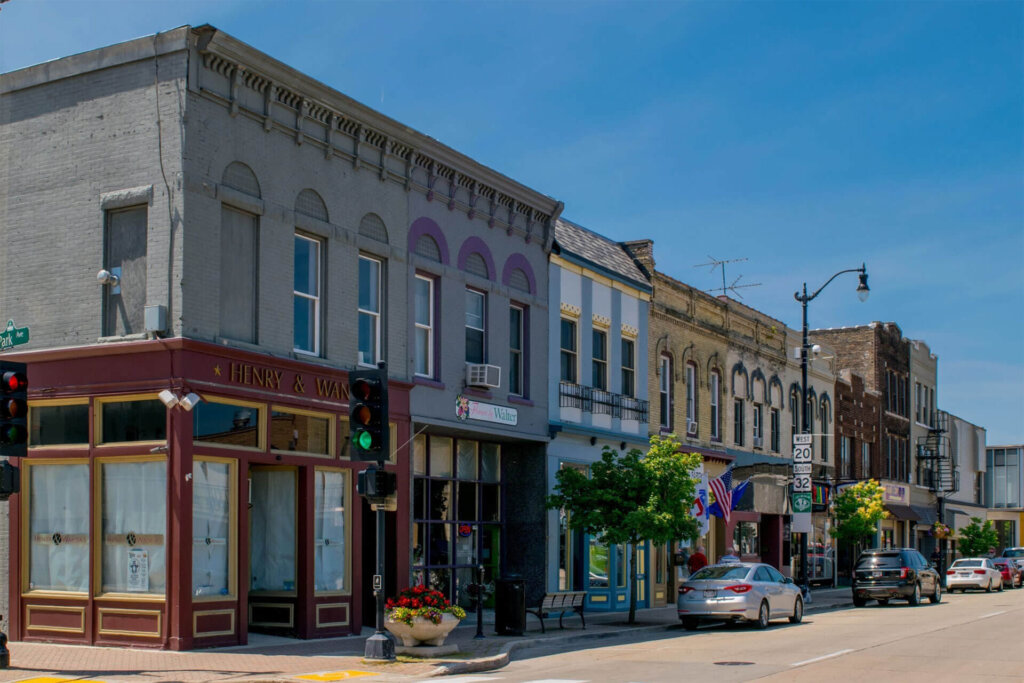 Historic downtown street in Brookfield, WI, featuring colorful storefronts, parked cars, and a clear blue sky.