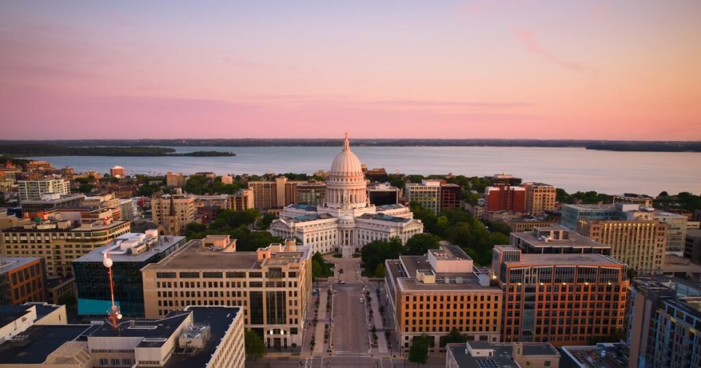 Aerial view of downtown Madison, Wisconsin at sunset, featuring the Wisconsin State Capitol building and surrounding cityscape near Lake Monona.
