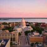Aerial view of downtown Madison, Wisconsin at sunset, featuring the Wisconsin State Capitol building and surrounding cityscape near Lake Monona.