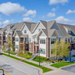 Modern multi-family apartment complex with gabled roofs, brick and siding exterior, located on a clean suburban street under a bright blue sky.