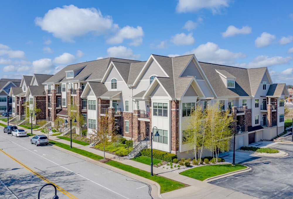 Modern multi-family apartment complex with gabled roofs, brick and siding exterior, located on a clean suburban street under a bright blue sky.