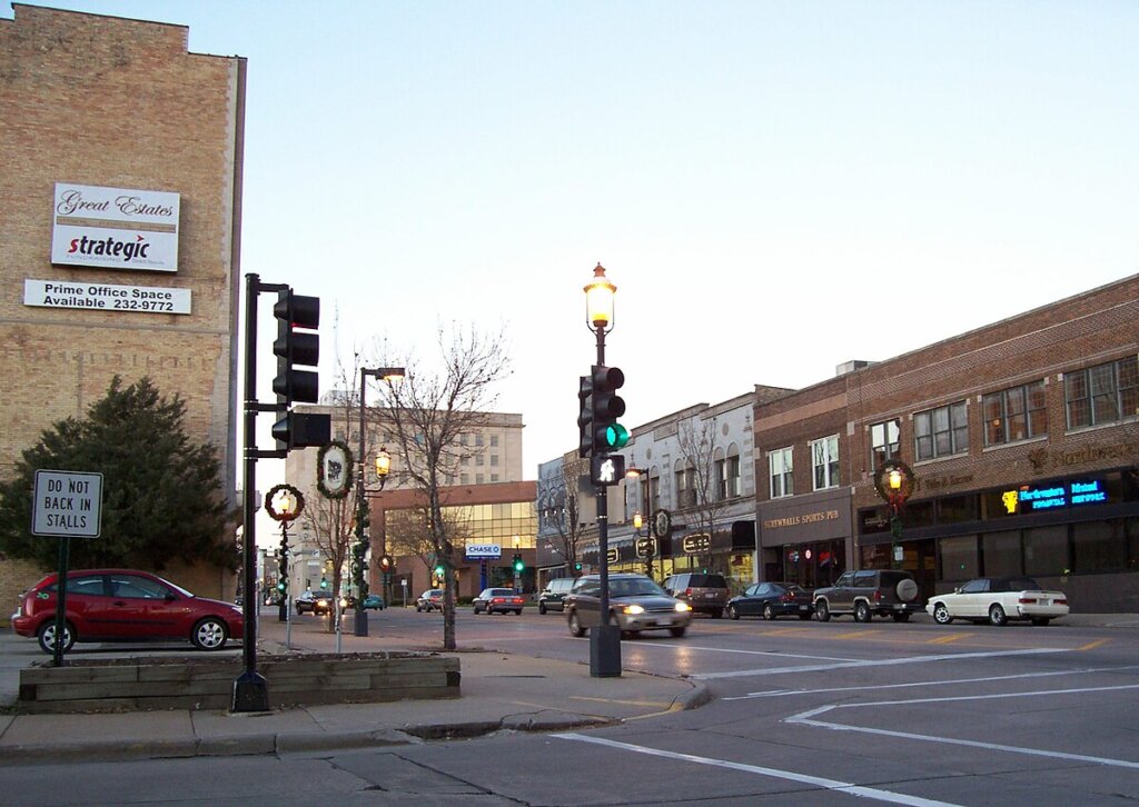 Downtown Oshkosh, Wisconsin, showing a busy street corner with historic brick buildings, storefronts, and traffic lights at dusk.