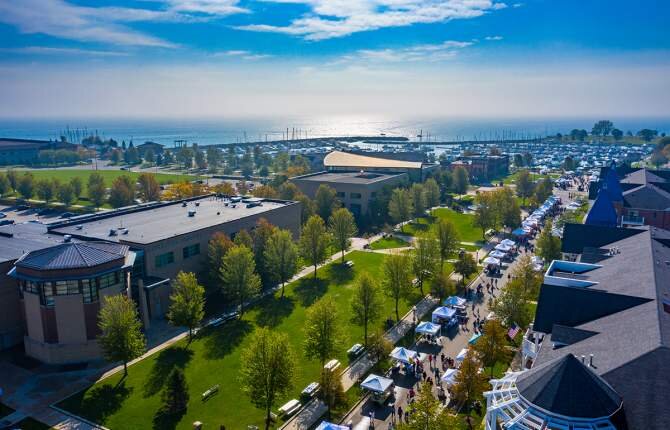 Aerial view of a lively outdoor market at HarborPark in Kenosha, with green lawns, tree-lined paths, and Lake Michigan in the background.