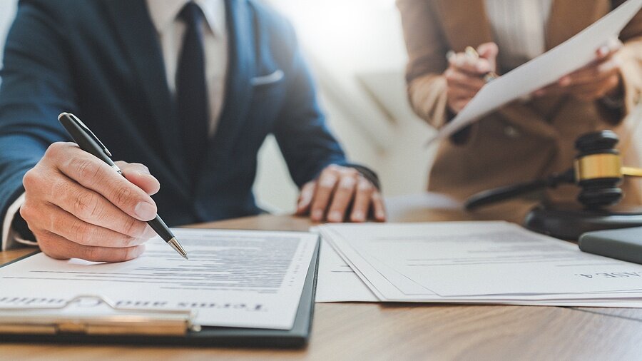 Close-up of a businessperson reviewing and signing legal documents at a desk, with another individual holding paperwork and a gavel nearby—illustrating the legal disclosure process when selling a house with water damage in Madison.