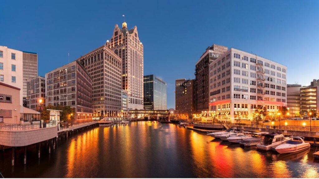 Downtown Milwaukee skyline at dusk reflecting on the river, with illuminated buildings and docked boats.