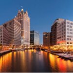 Downtown Milwaukee skyline at dusk reflecting on the river, with illuminated buildings and docked boats.