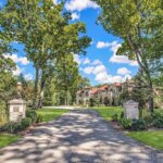Luxurious Brookfield estate with gated driveway, manicured lawn, and grand architecture surrounded by tall trees under a bright blue sky.
