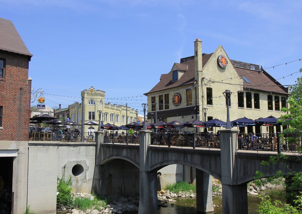 Scenic view of downtown Wauwatosa featuring a pedestrian bridge, outdoor dining, and historic buildings under a clear blue sky.