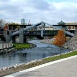 Scenic pedestrian bridge over a rocky river in downtown Waukesha, surrounded by trees and urban architecture.