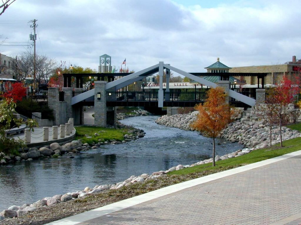 Scenic pedestrian bridge over a rocky river in downtown Waukesha, surrounded by trees and urban architecture.