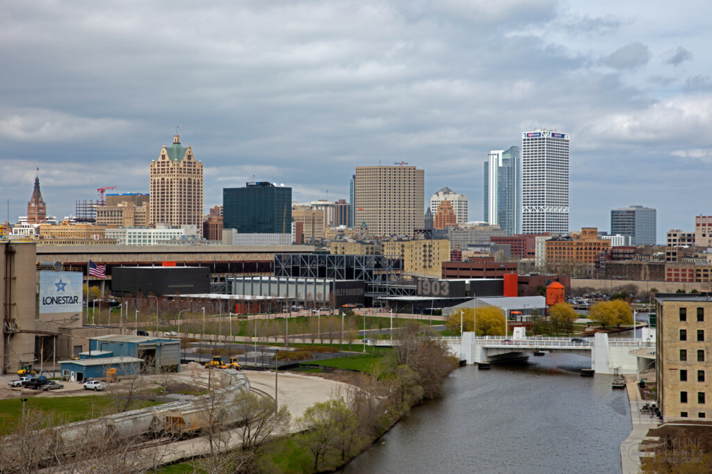 Downtown Milwaukee skyline featuring the Milwaukee River, modern high-rises, and the iconic Harley-Davidson Museum under a cloudy sky.