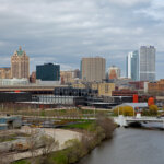 Downtown Milwaukee skyline featuring the Milwaukee River, modern high-rises, and the iconic Harley-Davidson Museum under a cloudy sky.