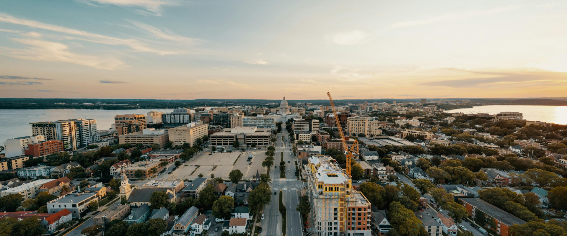 Aerial view of downtown Madison, Wisconsin at sunset, featuring the State Capitol and surrounding buildings, with lakes on both sides and ongoing construction in the foreground.