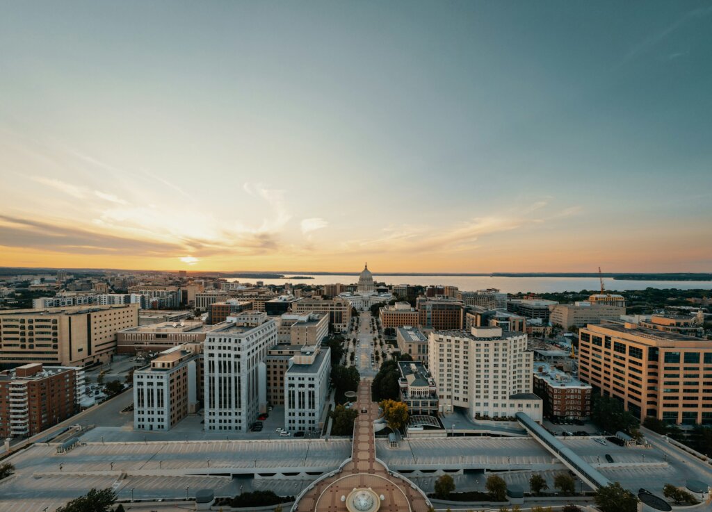 Aerial view of downtown Madison, Wisconsin at sunset, featuring the Wisconsin State Capitol and surrounding buildings with lakes in the background.