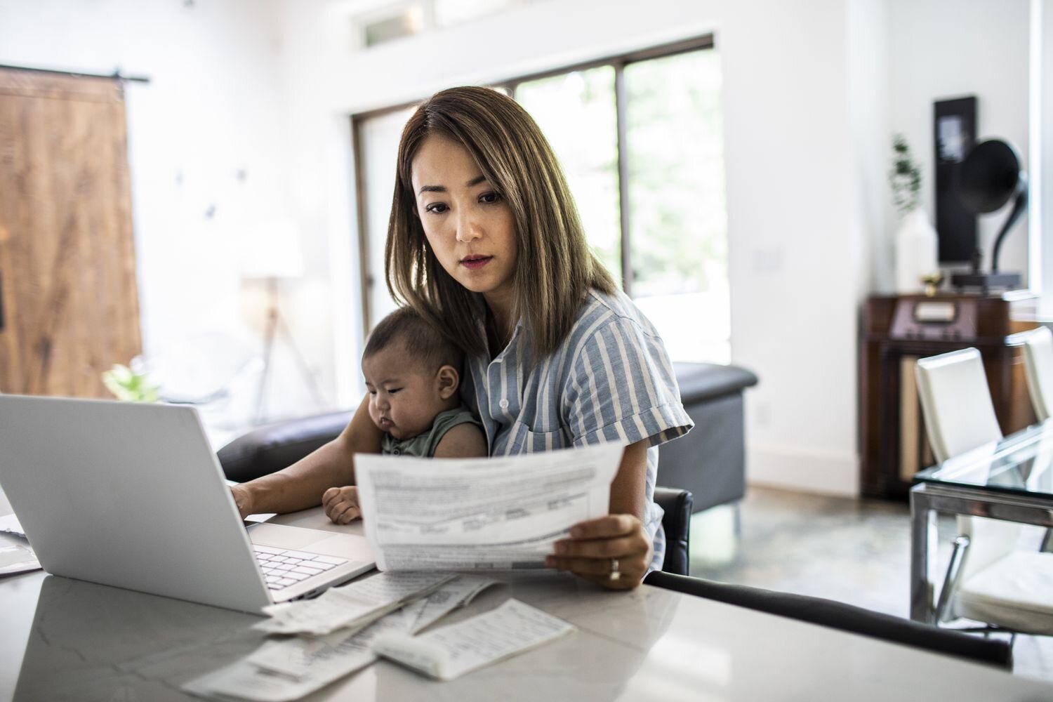 Worried mother holding her baby while reviewing documents at home with a laptop.