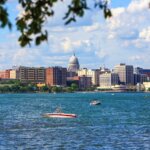 View of downtown Madison, Wisconsin with the State Capitol building, seen across Lake Monona with boats on the water and tree branches framing the scene.