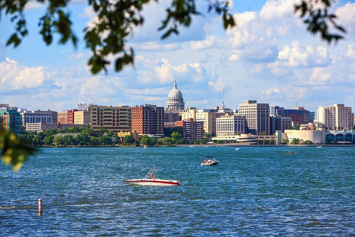 View of downtown Madison, Wisconsin with the State Capitol building, seen across Lake Monona with boats on the water and tree branches framing the scene.