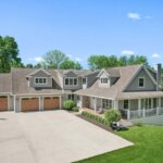 Spacious modern farmhouse with a three-car garage, wraparound porch, and manicured lawn on a sunny day.