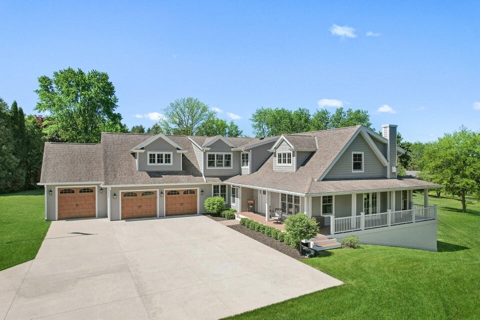 Spacious modern farmhouse with a three-car garage, wraparound porch, and manicured lawn on a sunny day.