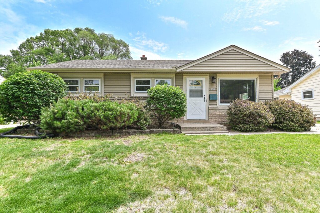 Single-story beige house in West Allis with trimmed bushes, a white front door, and a green lawn under a clear blue sky.