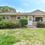 Single-story beige house in West Allis with trimmed bushes, a white front door, and a green lawn under a clear blue sky.
