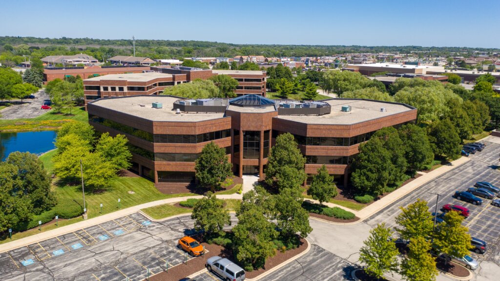 Aerial view of a modern commercial office building in Brookfield, Wisconsin, surrounded by trees, a pond, and a large parking lot on a clear summer day.