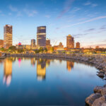 Downtown Milwaukee skyline at sunset with buildings reflecting on calm lake water and a rocky shoreline in the foreground.