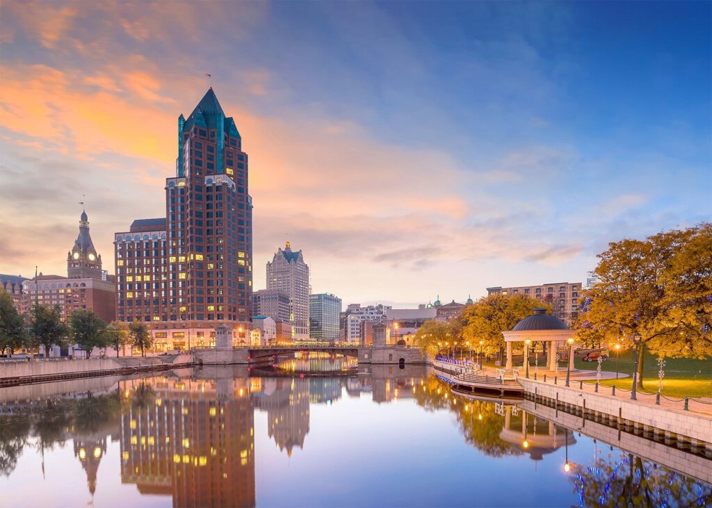 Downtown Milwaukee skyline at sunset with buildings reflecting on the Milwaukee River and autumn trees lining the Riverwalk.