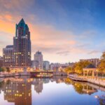 Downtown Milwaukee skyline at sunset with buildings reflecting on the Milwaukee River and autumn trees lining the Riverwalk.