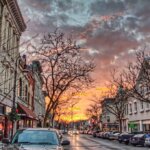 Downtown Kenosha street at sunset with historic buildings, parked cars, and leafless trees under a dramatic, colorful sky.