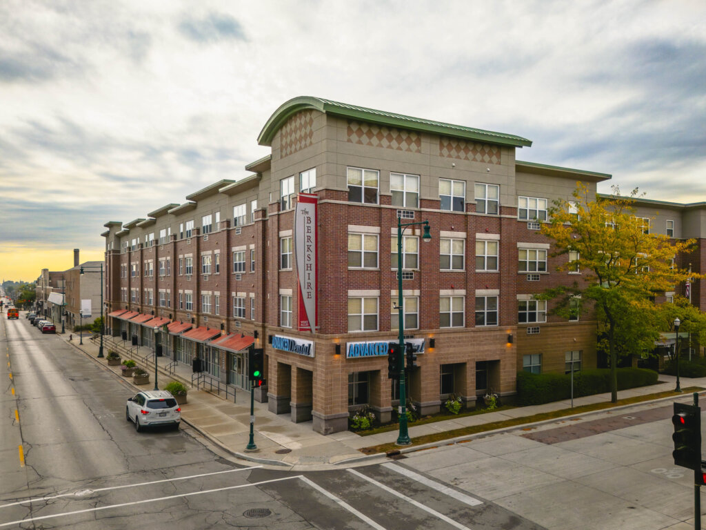 Street view of The Berkshire building in West Allis, a modern brick apartment complex with storefronts and tree-lined sidewalks under a cloudy sky.