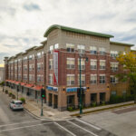 Street view of The Berkshire building in West Allis, a modern brick apartment complex with storefronts and tree-lined sidewalks under a cloudy sky.