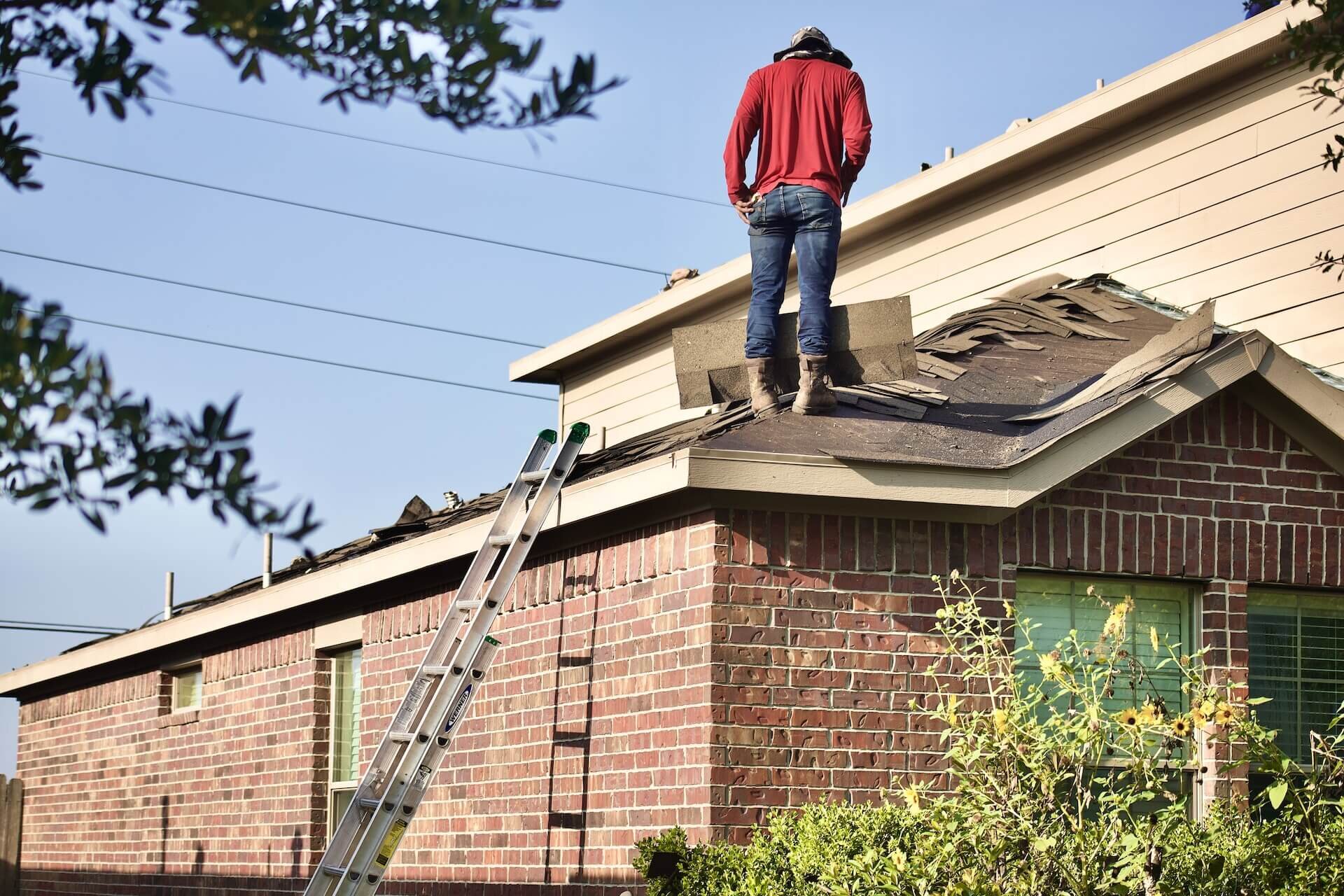 Worker repairing roof shingles on a brick house using a ladder.