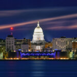 Nighttime view of downtown Madison, Wisconsin, featuring the illuminated Wisconsin State Capitol building reflected over Lake Monona.